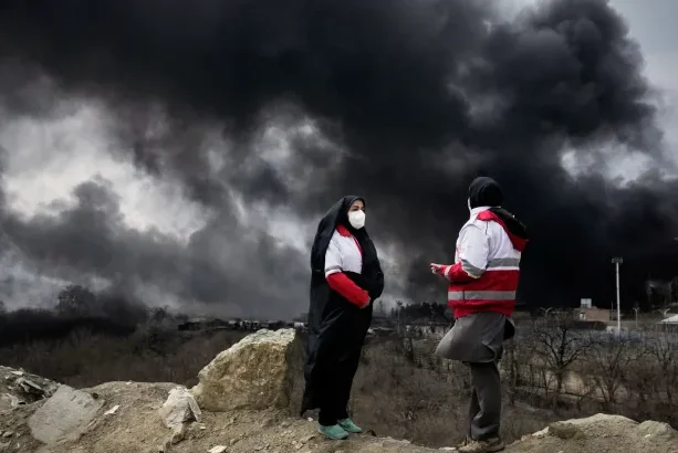 Two women from the Iranian Red Crescent Society stand as a thick plume of smoke from a United States-Israeli strike on an oil storage facility late Saturday rises in the sky in Tehran, Iran, Sunday, March 8, 2026 [Vahid Salemi/AP Photo] /Al Jajeera